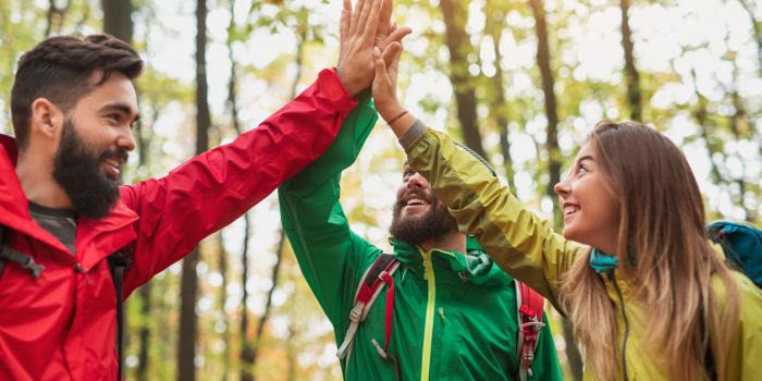 Group of people in colorful outerwear smiling and giving high five while traveling through forest in autumn.